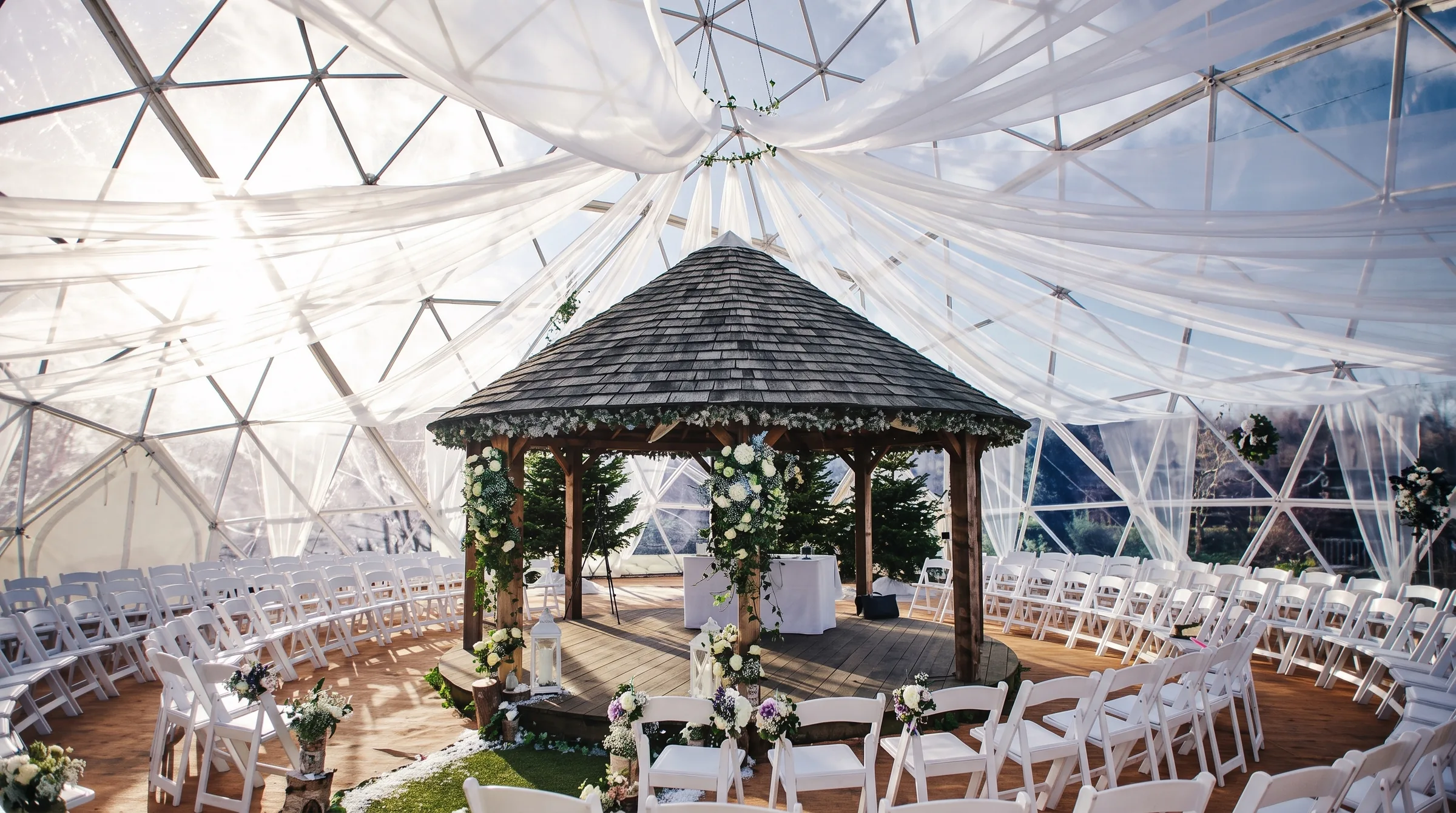 Wedding ceremony set in the round under a clear geodesic dome, with white draping and a wooden gazebo